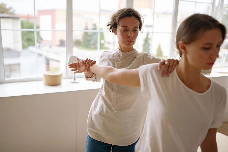 A therapist guides a patient's outstretched arm through a shoulder stretch by a sunny window